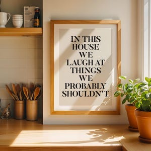 Puede incluir: Un marco de madera con el texto "IN THIS HOUSE WE LAUGH AT THINGS WE PROBABLY SHOULDN'T" se encuentra sobre una encimera de madera. Dos plantas en macetas est&aacute;n a la derecha del marco. Utensilios de cocina y decoraci&oacute;n visibles.