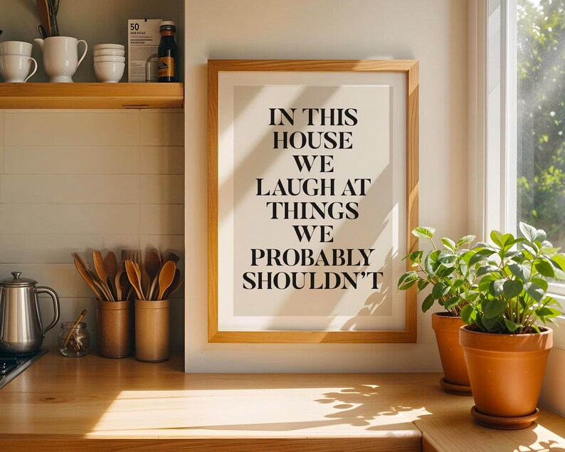 Puede incluir: Un marco de madera con el texto "IN THIS HOUSE WE LAUGH AT THINGS WE PROBABLY SHOULDN'T" se encuentra sobre una encimera de madera. Dos plantas en macetas est&aacute;n a la derecha del marco. Utensilios de cocina y decoraci&oacute;n visibles.