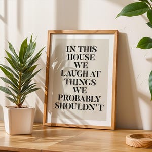 Puede incluir: Un cuadro enmarcado con el texto "IN THIS HOUSE WE LAUGH AT THINGS WE PROBABLY SHOULDN'T" en fuente negra. El cuadro est&aacute; en un marco de madera clara y se encuentra sobre una superficie de madera. Una planta en maceta est&aacute; a la izquierda.