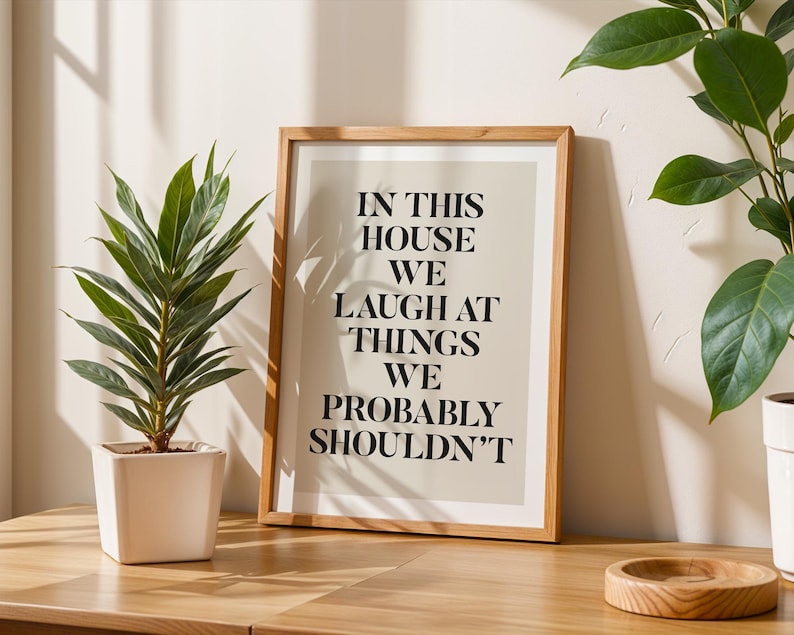 Puede incluir: Un cuadro enmarcado con el texto "IN THIS HOUSE WE LAUGH AT THINGS WE PROBABLY SHOULDN'T" en fuente negra. El cuadro est&aacute; en un marco de madera clara y se encuentra sobre una superficie de madera. Una planta en maceta est&aacute; a la izquierda.
