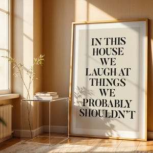 Puede incluir: Un cuadro enmarcado con el texto "IN THIS HOUSE WE LAUGH AT THINGS WE PROBABLY SHOULDN'T" en fuente negra. La impresi&oacute;n est&aacute; en un marco dorado y se apoya contra una pared beige. Una mesa auxiliar transparente con libros y una planta est&aacute; frente a una ventana.