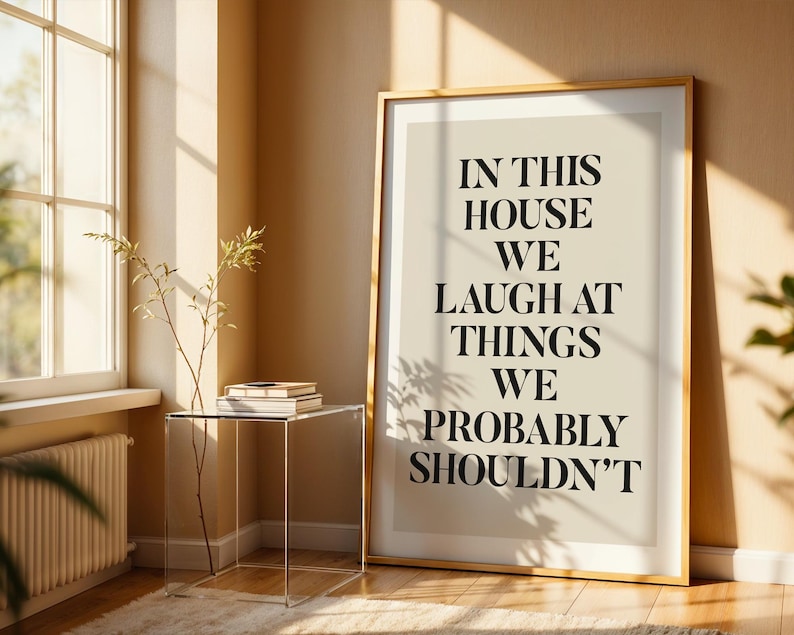 Puede incluir: Un cuadro enmarcado con el texto "IN THIS HOUSE WE LAUGH AT THINGS WE PROBABLY SHOULDN'T" en fuente negra. La impresi&oacute;n est&aacute; en un marco dorado y se apoya contra una pared beige. Una mesa auxiliar transparente con libros y una planta est&aacute; frente a una ventana.