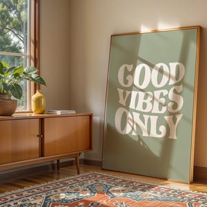 Op de afbeelding: Een ingelijste poster met de woorden "GOOD VIBES ONLY" in het wit, leunend tegen een muur. De poster staat in een kamer met een houten kast, een plant en een patroonkleed. De poster heeft een groene achtergrond en een gouden lijst.
