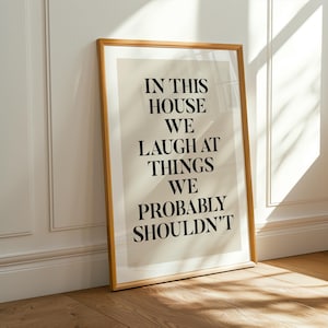 Puede incluir: Un cuadro enmarcado con el texto "IN THIS HOUSE WE LAUGH AT THINGS WE PROBABLY SHOULDN'T" en letras negras. El cuadro tiene un fondo beige claro y un marco de madera. Se apoya contra una pared blanca con luz solar.