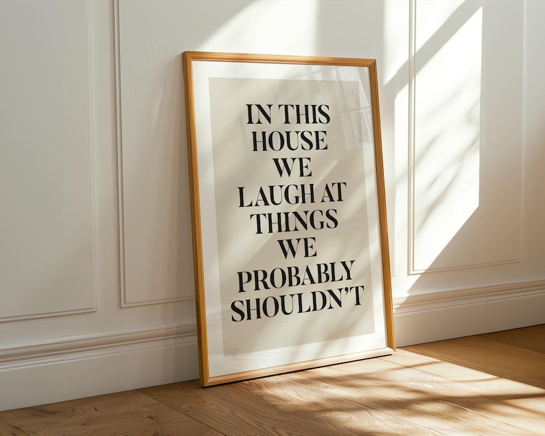 Puede incluir: Un cuadro enmarcado con el texto "IN THIS HOUSE WE LAUGH AT THINGS WE PROBABLY SHOULDN'T" en letras negras. El cuadro tiene un fondo beige claro y un marco de madera. Se apoya contra una pared blanca con luz solar.