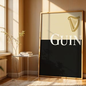 May include: A framed poster featuring the Guinness logo. The top half is a light beige color, and the bottom half is black. The word "Guinness" is written in white, bold letters. A gold harp logo is in the top right corner.