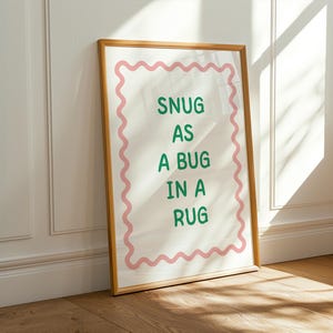 Peut inclure: Une affiche encadrée avec les mots "SNUG AS A BUG IN A RUG" en vert. Le texte est centré sur un fond blanc, encadré par une bordure rose ondulée. Le cadre est de couleur marron clair, et l'affiche est appuyée contre un mur blanc.