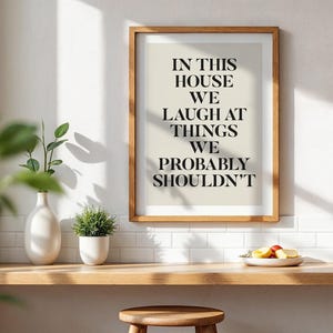 Puede incluir: Una impresi&oacute;n enmarcada con el texto "IN THIS HOUSE WE LAUGH AT THINGS WE PROBABLY SHOULDN'T" en fuente negra. La impresi&oacute;n est&aacute; en un marco de madera y se exhibe en un estante de madera con un jarr&oacute;n blanco, una peque&ntilde;a planta en maceta y un plato de fruta.