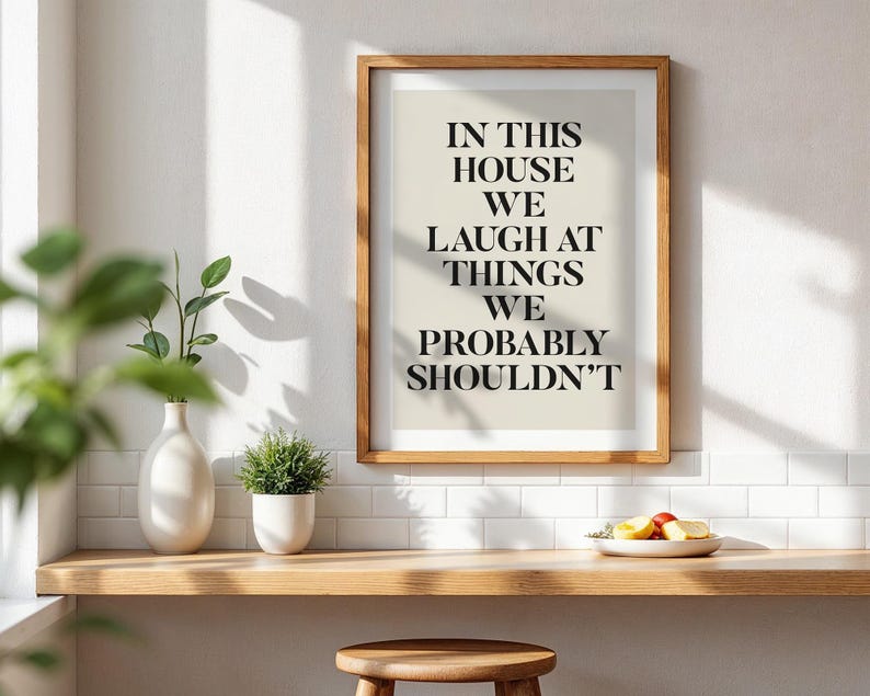 Puede incluir: Una impresi&oacute;n enmarcada con el texto "IN THIS HOUSE WE LAUGH AT THINGS WE PROBABLY SHOULDN'T" en fuente negra. La impresi&oacute;n est&aacute; en un marco de madera y se exhibe en un estante de madera con un jarr&oacute;n blanco, una peque&ntilde;a planta en maceta y un plato de fruta.