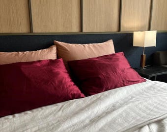 Puede incluir: Una cama con un edredón blanco a rayas y varias almohadas. Dos almohadas de color rojo oscuro están en primer plano, con dos almohadas de color melocotón detrás. El cabecero es azul oscuro con paneles de madera encima.