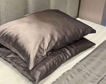 Puede incluir: Dos fundas de almohada grises de satén apiladas sobre una sábana blanca a rayas. Las fundas de almohada tienen un brillo lustroso y son de color liso y neutro. La ropa de cama es parte de un juego de dormitorio.