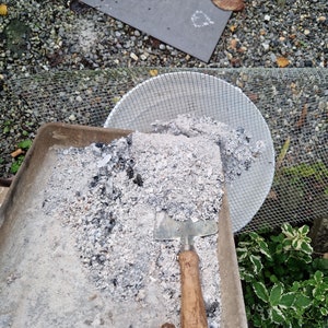 May include: A metal tray filled with white ash and a small gardening trowel. The tray is sitting on top of a wire mesh screen.
