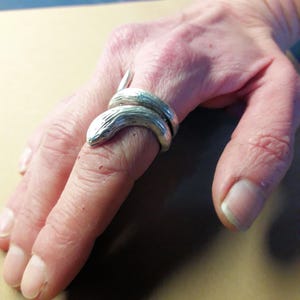 May include: A silver-colored ring in the shape of a snake coiled around a finger. The snake's head is detailed with scales and a small eye. The ring appears to be made of metal and is worn on a person's finger.