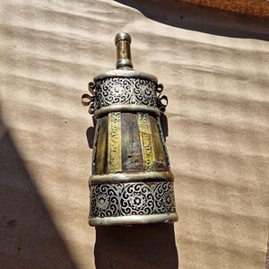 Könnte beinhalten: Eine antike Flasche mit silbernem Filigranmuster an Ober- und Unterseite. Der Flaschenkörper ist eine Mischung aus Braun, Grün und Gold. Die Flasche hat einen silbernen Deckel und kleine Griffe an jeder Seite.