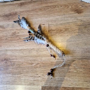 Puede incluir: Una varita de plumas hecha a mano con un mango de cuerda envuelta y cuentas de madera. La varita presenta un grupo de plumas marrones y blancas en la parte superior y está diseñada con materiales naturales. La varita está sobre una superficie de madera.