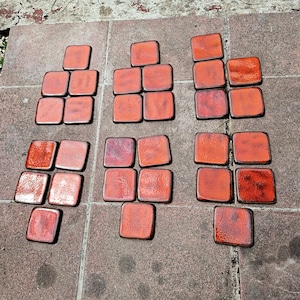 Op de afbeelding: Een verzameling vierkante keramische onderzetters in tinten oranje en rood. De onderzetters zijn in groepen gerangschikt en tonen hun glanzende afwerking en verschillende tinten. De onderzetters zijn ongeveer 10 cm vierkant.