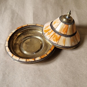 May include: A round brass dish with a matching conical lid, both featuring inlaid segments of cream and orange-toned material. The metal edges are decorated with a twisted rope pattern, and the lid is topped with a small, pointed brass finial.