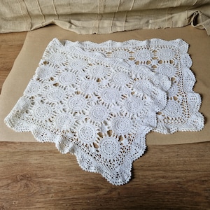 May include: A white, handmade crochet doily with a floral pattern. The doily is square-shaped with a scalloped edge. The intricate design features circular motifs connected by delicate lacework. The doily is displayed on a brown surface.