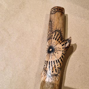 May include: A wooden stick with intricate black ink designs. The artwork includes a central starburst pattern and other geometric shapes. The stick is a natural light brown color, with the designs adding contrast.