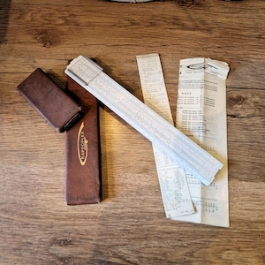 Può includere: Un regolo calcolatore vintage con custodia in pelle marrone e documenti. Il regolo calcolatore è bianco e blu con segni di misurazione. La custodia è impressa con la scritta "RAPHOPLEX". I documenti includono testo e calcoli.