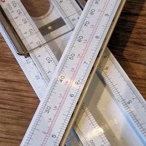 Może przedstawiać: Widok z góry na kilka białych i szarych narzędzi pomiarowych, w tym linijki i kątomierz, zachodzących na siebie na drewnianej powierzchni. Linijki wyświetlają pomiary w centymetrach i calach. Narzędzia są ułożone pod różnymi kątami.