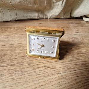 Op de afbeelding: Een vintage goudkleurige reiswekker met een lichtgekleurde wijzerplaat en zwarte cijfers. De klok is rechthoekig met een scharnierende bovenkant en een bruine kunstleren hoes. Het woord "DELUXE" staat op de wijzerplaat.