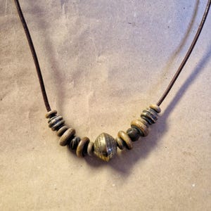 Può includere: Una collana artigianale con un cordino in pelle marrone e una serie di perline in legno e metallo. Il punto focale è una perlina metallica strutturata, che aggiunge un'estetica rustica e terrosa. Per uno stile naturale e bohémien.
