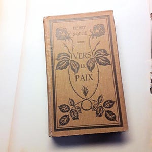 Peut inclure: Un livre ancien avec une couverture beige. Le titre "VERS LA PAIX" est écrit en noir, ainsi que le nom de l'auteur, "HENRY SOULIE". La couverture est ornée d'illustrations florales et de feuilles noires.