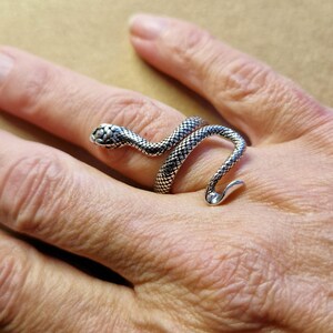 Op de afbeelding: Een zilverkleurige slangenring gewikkeld om een vinger. De ring heeft een gedetailleerde slangenkop en -lichaam met een textuurpatroon. De staart van de slang krult om de vinger, wat een uniek en opvallend ontwerp creëert.