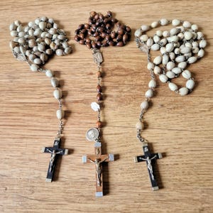 Può includere: Tre rosari con diversi colori di perline e disegni di croci. Un rosario ha perline marrone scuro, un altro perline bianco sporco e il terzo perline di colore chiaro. Le croci sono nere e argentate o in legno con una figura argentata.