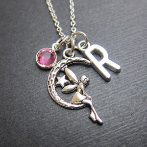 Puede incluir: Collar de plata con un dije de cristal rosa, un dije de luna creciente con un hada sentada en él y un dije de la letra "R".