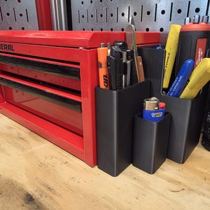 May include: A red US General toolbox with three drawers, next to black organizers holding tools and a blue Bic lighter. Various tools are visible, including a Milwaukee tool and a can labeled "Model 72 Flash Bang".
