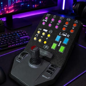 Op de afbeelding: Een zwarte gaming-controller met een centrale joystick, een gestructureerd bedieningspaneel met diverse gekleurde knoppen en labels zoals ENGINE START, JOB INFO en VEHICLE INFO. Het apparaat bevat blauwe, groene, gele en rode knoppen.