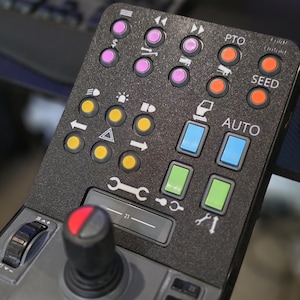 Könnte beinhalten: Eine schwarze Steuerung mit mehreren Knöpfen und einem Joystick. Das Panel verfügt über Knöpfe in verschiedenen Farben, darunter Pink, Orange, Gelb, Blau und Grün, mit Beschriftungen wie "PTO", "SEED" und "AUTO".