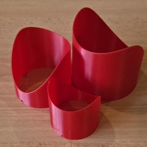May include: Three red, curved, open-topped containers of varying sizes are arranged on a light brown wooden surface. The containers have a smooth, glossy finish and a rounded, abstract shape, suggesting a modern design.