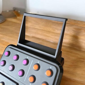 May include: Black music production controller with a built-in screen and multiple coloured buttons. The controller has a black frame and a wooden surface.