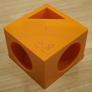 Puede incluir: Cubo de color naranja brillante con una abertura triangular en la parte superior y dos aberturas circulares en los lados. El texto "2.5" 1.0D" está impreso en el lateral. El cubo es de material sólido.