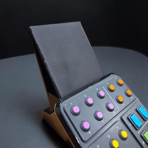 Pode incluir: Um suporte inclinado preto segurando um painel de controle cinza com botões numerados. O painel apresenta fileiras de botões circulares em roxo, laranja, amarelo e azul, dispostos em uma grade com pequenos números brancos impressos.