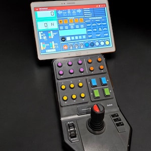 Puede incluir: Un panel de control negro con una palanca de mando y una variedad de botones. El panel está conectado a una tableta Samsung que muestra una interfaz de control con el texto "ON" y "STATUS".