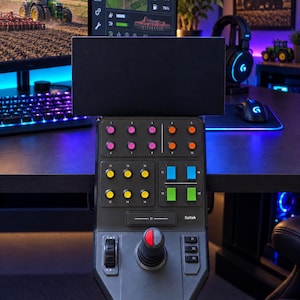 May include: A black Saitek gaming control panel for simulation games, featuring a central joystick, a horizontal slider, and a grid of numbered buttons in pink, orange, yellow, blue, and green, mounted to the edge of a dark computer desk.