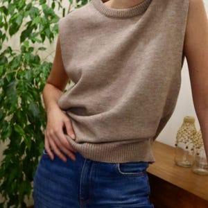 Op de afbeelding: Een bruine, mouwloze gebreide trui met een ronde hals. De trui wordt gedragen over een blauwe jeans.