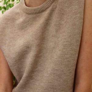 Op de afbeelding: Een mouwloze gebreide trui in een licht beige kleur. De trui heeft een ronde hals en een losse pasvorm.