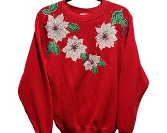 May include: A red sweatshirt with white poinsettia flower and green leaf embroidery. The long-sleeved top is hanging on a black hanger. The floral design is on the left shoulder and across the chest.