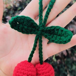 Op de afbeelding: Een handgemaakt gehaakt ornament met twee felrode bolvormige kersen bevestigd aan groene steeltjes en twee groene blaadjes. De kersen zijn aan de bovenkant verbonden door een groen gehaakt lusje, ontworpen als decoratief hangend accessoire.