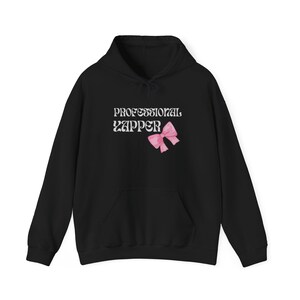 Puede incluir: Sudadera con capucha negra con el texto "PROFESSIONAL YAPPER" en blanco y un lazo rosa debajo.