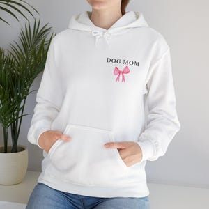 Puede incluir: Sudadera con capucha blanca con un bolsillo grande y el texto "DOG MOM" impreso en la parte delantera. Un lazo rosa está centrado debajo del texto.