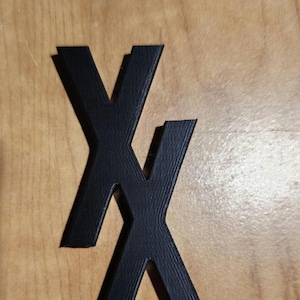May include: Two black plastic letters, each shaped like the letter "X", are stacked on top of each other. The letters are on a light brown wooden surface.