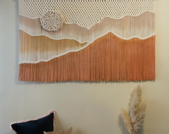 May include: A macrame wall hanging with a peach and cream color scheme. The hanging features a large circle in the center and a wave-like design. The hanging is made with natural fibers and has a rustic, bohemian aesthetic.
