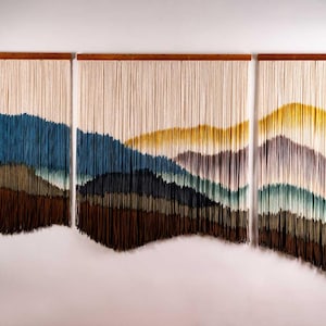 Op de afbeelding: Een wandkleed gemaakt van garen met een landschapsontwerp. Het kleed is verdeeld in drie secties, elk met een ander kleurenschema. De linker sectie is blauw, groen en bruin. De middelste sectie is blauw, groen en geel. De rechter sectie is paars, groen en geel.