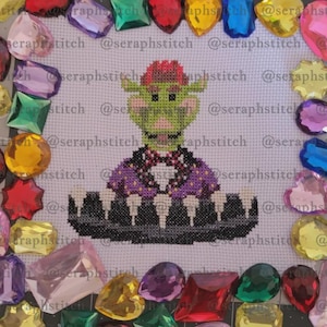 May include: Cross-stitch of a green goblin-like creature with a red hair, wearing a purple and black outfit. The artwork is surrounded by colorful, gem-like embellishments. The text "@seraphstitch" is visible.